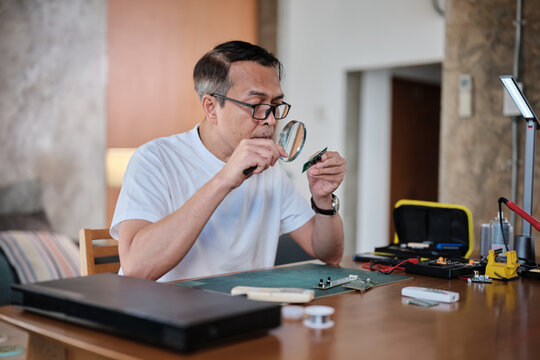 Mature Asian man looks at and considers circuit board details with magnifying glass, focusing on precision checks for home electrical appliance DIY fix, electronics component hobby, handyman workshop.