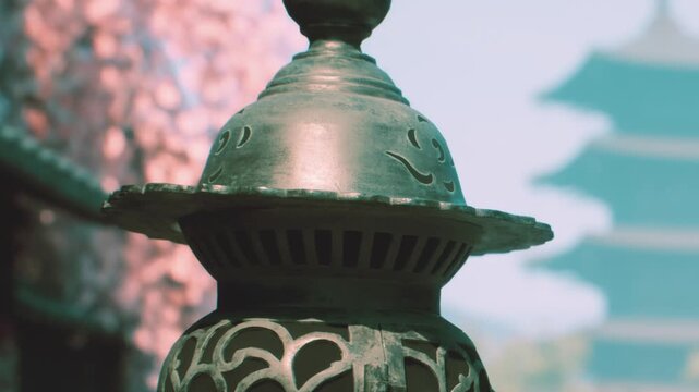 A metal lantern stands in a temple courtyard in Japan. Its detailed design is visible against a clear sky with pagodas in the background. People walk around the area.
