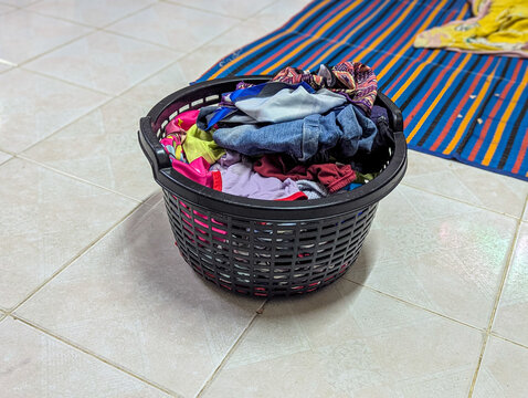 Black plastic laundry basket filled with clothes on tiled floor.