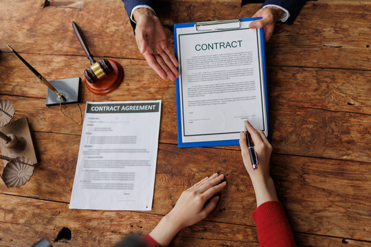 Person signs a formal contract on a clipboard at a rustic wooden desk with a legal professional nearby, finalizing a business agreement, signature and paperwork in view