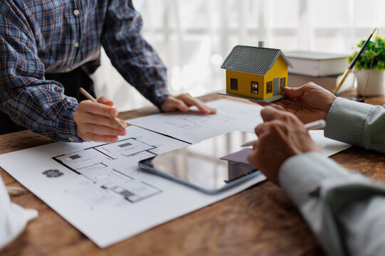 Real estate agents and clients review architectural blueprints and a house model around a tablet, discussing contracts, financing and project planning for a new home purchase