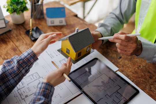 Architect and client discussing property investment, reviewing house model, blueprint plans, and digital tablet, planning construction and real estate project