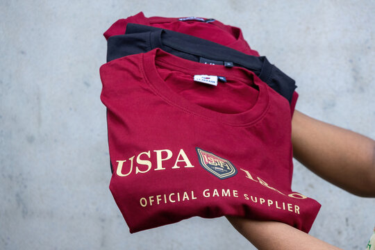 Kishoreganj, Bangladesh - 20 August, 2026 - Close-up of human hands holding stack of folded branded t shirts. Red USPA printed casual apparel presented outdoors.