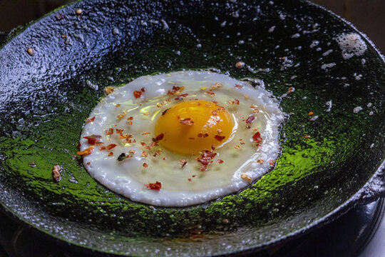 Freshly Cooked Sunny Side Up Fried Egg Sprinkled with Red Chili Flakes in Hot Frying Pan, Close Up View of Egg Yolk, Homemade Healthy Breakfast Meal.