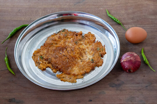 Crispy Fried Egg Omelet Cooked with Onions and Green Chilies, Served on Stainless Steel Plate with Whole Egg, Onion, Green Chili Props, Wooden Background.