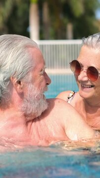 Cute senior couple relaxing and enjoying the swimming pool together on a sunny day. Romantic retired pensioners having fun on vacation, hugging, kissing and laughing in the water