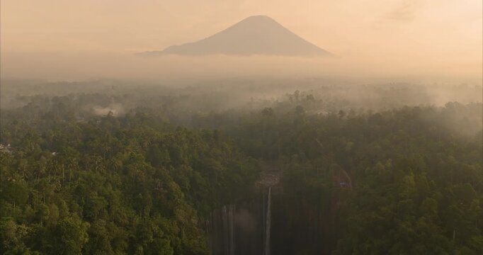 Aerial view of Tumpak Sewu Waterfall surrounded by lush tropical rainforest and mist with a distant volcano peak at sunrise in Kecamatan Pronojiwo, Jawa Timur, Indonesia.