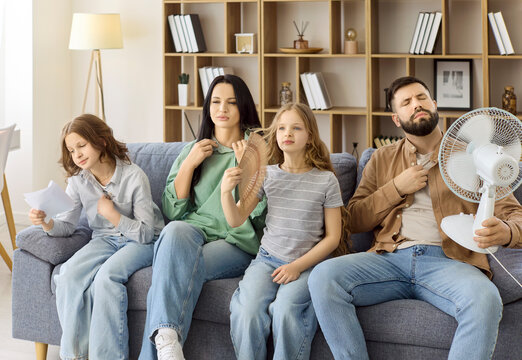 Family with parents and children in hot home. They sit on a couch, in sweat and fanning for relief as a fan blows, showing summer heat, discomfort, and no cooling. Concept of overheating at home