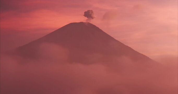Aerial view of a volcanic peak emitting smoke above a sea of clouds during a vibrant sunset in Kecamatan Pronojiwo, Jawa Timur, Indonesia.