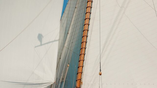 Shadow of person perched high in the mast against billowing white sails overhead