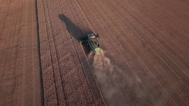 An aerial view of a green combine harvester harvesting crops in a dusty field in Norway, with a long shadow cast by the low sun