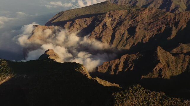 Aerial view of rugged volcanic mountain peaks in Tenerife with clouds filling the deep valleys under golden hour light in Santa Cruz de Tenerife, Canary Islands, Spain.