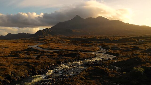 Aerial view of the rugged Skye landscape with a winding river flowing through the moorland under a golden sunset sky in Highland Council, Scotland, United Kingdom.
