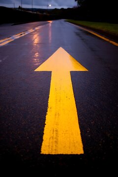 Bright yellow directional arrow painted on wet asphalt road surface during twilight