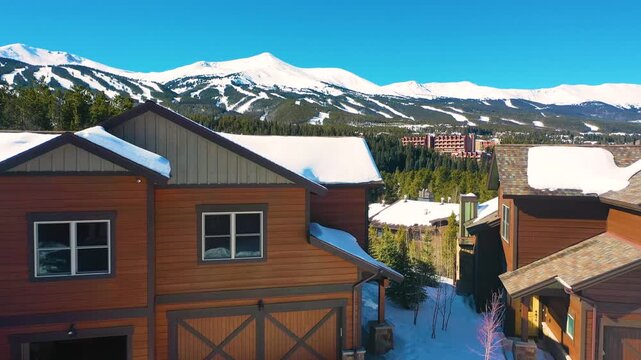 Aerial view of luxury mountain townhomes with snow covered roofs and scenic snowy peaks in background during sunny winter day.