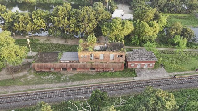 Drone lateral move past abandoned two-storey red railside quarters beside single track and ponds in rural Bangladesh