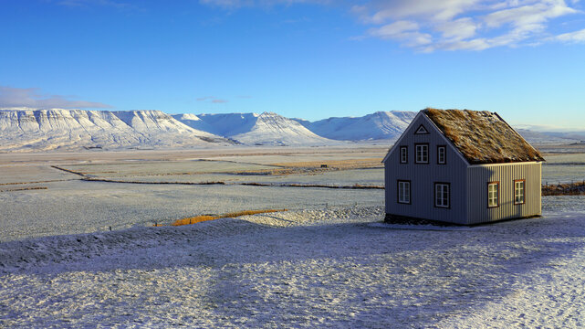 A traditional turf-roof house stands on a snow-covered hill overlooking a vast winter plain and distant snow-capped mountains in Iceland