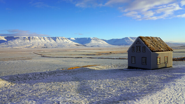 A traditional turf-roofed house stands on a snow-covered rise overlooking an expansive winter plain and distant snow-capped mountain range under a blue sky in Iceland
