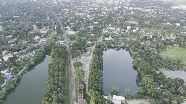 Overhead drone push along tree strip rail corridor with twin ponds and dense settlement in Bangladesh