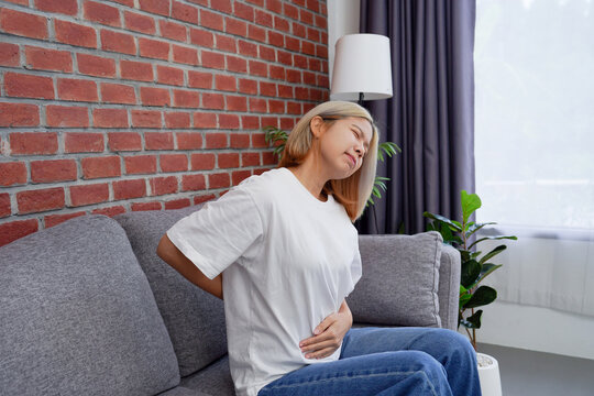 Portrait of Young asian woman has waist pain or lumbago and massaging her while sitting on sofa in the living room, Concepts of health problems