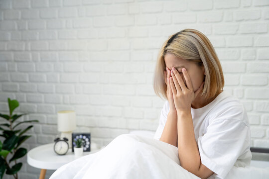 Stressed Asian woman sits on the bed, covering her face with her hands.