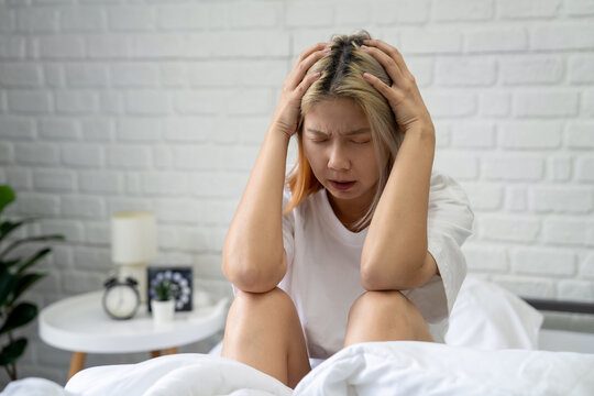 Stressed Asian woman sits on the bed, placing her free hand on her head unhappy female spending early morning in bedroom at home