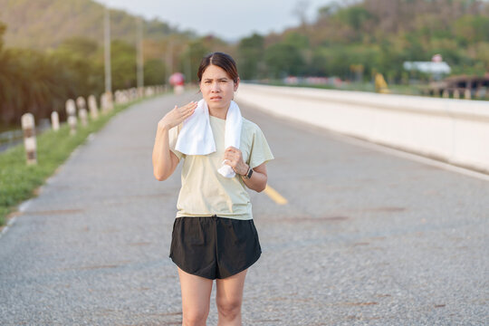 Exhausted Asian woman wiping sweat with a towel after jogging in the park. Tired female runner suffering from summer heat and fatigue after fitness workout. Outdoor exercise and heatstroke concept