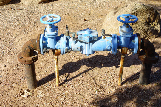 Blue painted water backflow preventer assembly with two handwheel shutoff valves and rusty plumbing pipes on ground outdoors