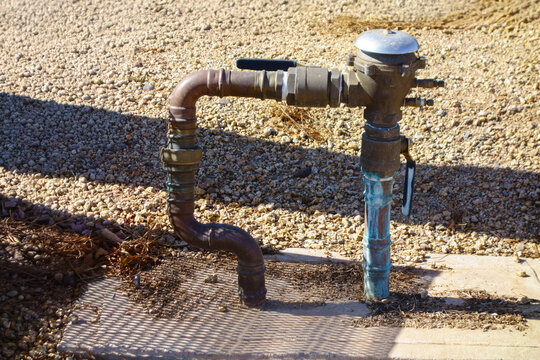 Outdoor brass pressure vacuum breaker backflow preventer assembly unit connected to weathered copper water plumbing pipe lines over gravel ground on sunny day.
