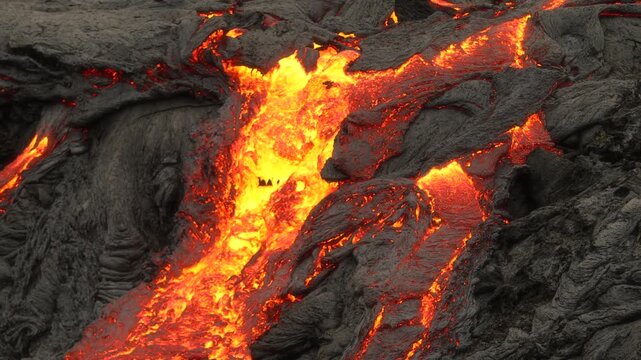 Aerial view of bright orange molten lava flowing steadily over dark, solidified volcanic rock formations in Iceland.