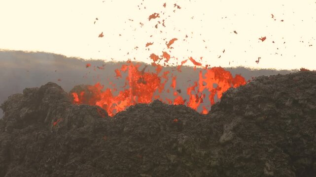 Aerial view of a volcanic eruption with molten lava splashing from a dark crater against a bright sky in Iceland.