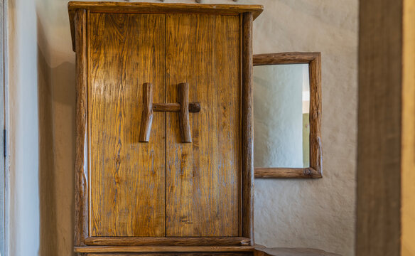 Rustic-style wardrobe. The door flaps and deadbolt are made of unpainted natural wood. There's a framed mirror on the rough plastered wall. Hotel