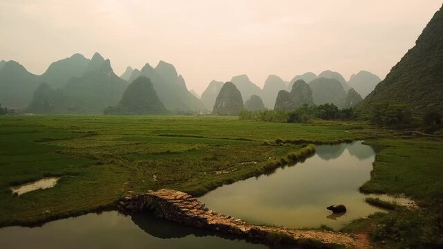 Aerial view of the iconic karst mountains and lush green rice paddies surrounding a small stone bridge over a calm river in Guilin, Guangxi, China.