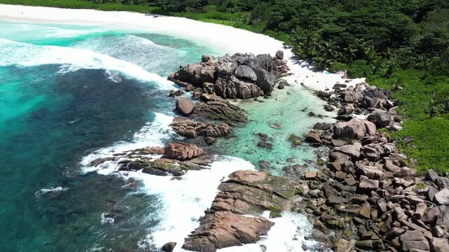 Aerial view of turquoise ocean waves crashing against massive granite boulders on tropical beach in La Digue Seychelles.