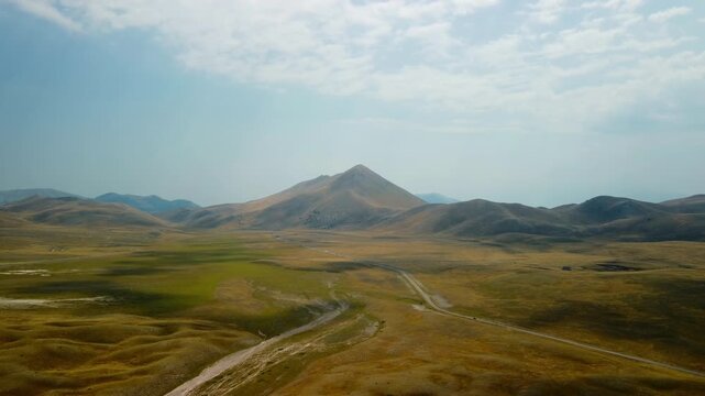 Drone mountain road golden plateau, aerial footage of vast grassy valley with winding highway leading to pyramid shaped peak under dramatic cloudy sky.