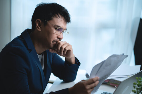 Stressed Businessman Reviewing Documents and Working on Laptop at Office Desk, Analyzing Business Problems and Financial Pressure