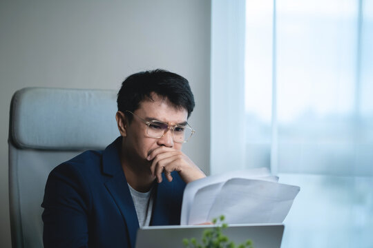 Businessman reviewing documents while working on a laptop in a modern office, showing concern and stress while analyzing business information, financial problems, 