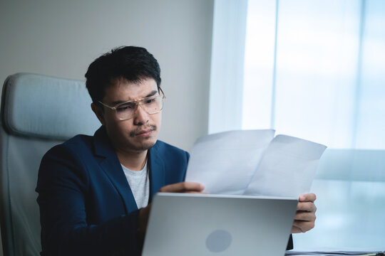 Businessman reviewing documents while working on a laptop in a modern office, showing concern and stress while analyzing business information, financial problems, workload, deadlines, 