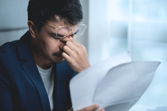 Stressed Businessman Reading Financial Documents While Working on Laptop at Office, Representing Business Crisis, Financial Pressure and Difficult Decisions