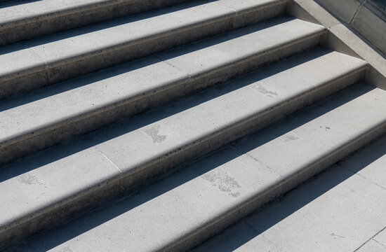 Close-up of wide stone stairs with strong diagonal sunlight and shadows, showing geometric lines, textured surfaces and footprints on urban outdoor steps.