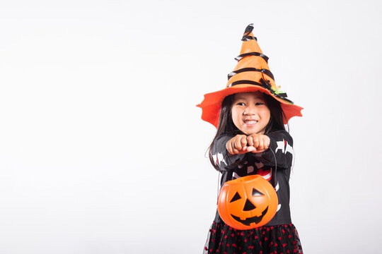 Halloween celebration showing cute kid wearing witch costume and funny hat while holding orange pumpkin bucket for trick or treat candy collection against empty isolated white studio backdrop