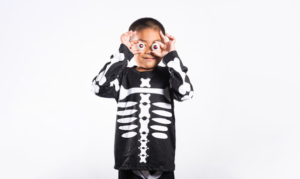Halloween celebration featuring kid wearing skeleton costume holding fake eyeballs over eyes on isolated white background