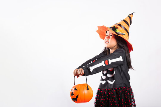 Halloween celebration featuring kid wearing witch hat and skeleton costume holding pumpkin bucket while standing against white isolated background ready for trick or treat fun activity