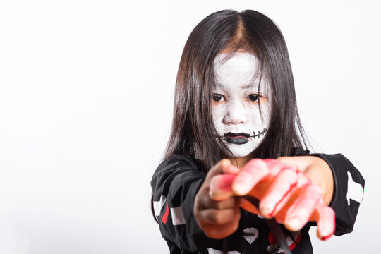 Halloween kid wearing white face paint and black costume holding fake bloody hand prop while smiling happily for autumn seasonal celebration party portrait against plain studio isolated background