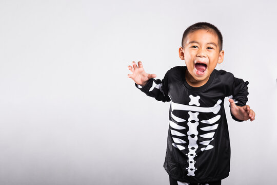 Halloween kid wearing skeleton costume playing spooky role while shouting joyfully during celebration event in studio with bright white isolated background lighting