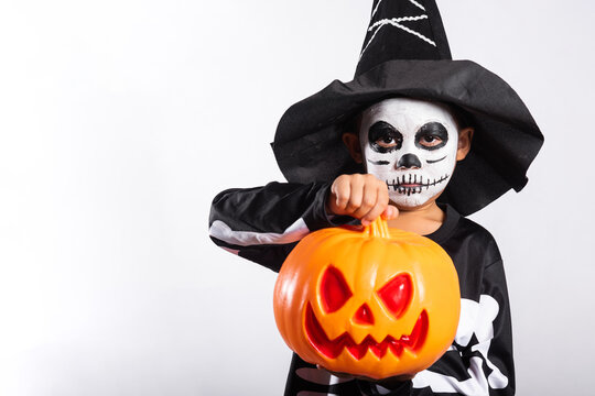 Halloween kid holding orange plastic pumpkin bucket wearing skeleton costume and witch hat celebrating spooky holiday festival wearing skull face paint smiling brightly white background studio