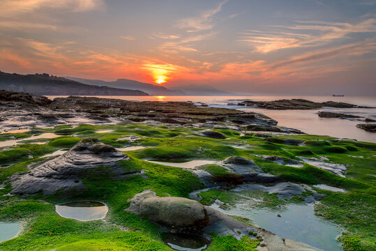 Golden sunset over rocky coast with vibrant green moss tide pools and dramatic sky at Lastron beach in Zierbena
