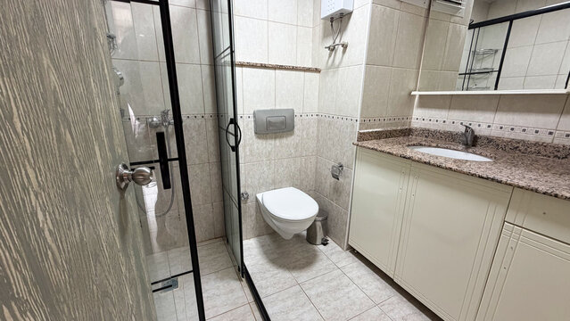 Compact residential bathroom interior with shower enclosure, wall mounted toilet, sink cabinet and tiled walls.