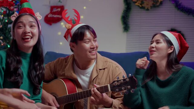 Happy asian friends singing christmas carols together playing guitar and giving gifts during a home party celebration