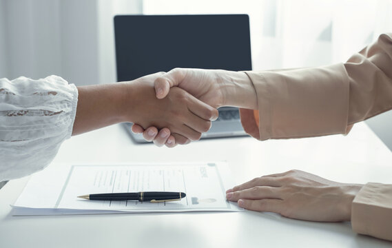 Business partners shaking hands over resume document and laptop at desk for job interview success hiring agreement and partnership concept